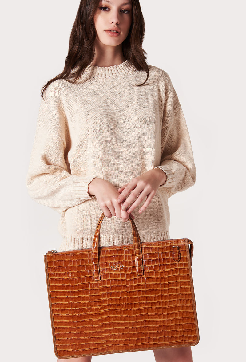 A model carries a sophisticated tan croco leather slim briefcase, showcasing its sophisticated design. The bag features a detachable shoulder strap, adding to its elegant appeal. The model confidently displays the bag's size and craftsmanship while exuding a sense of style and elegance.