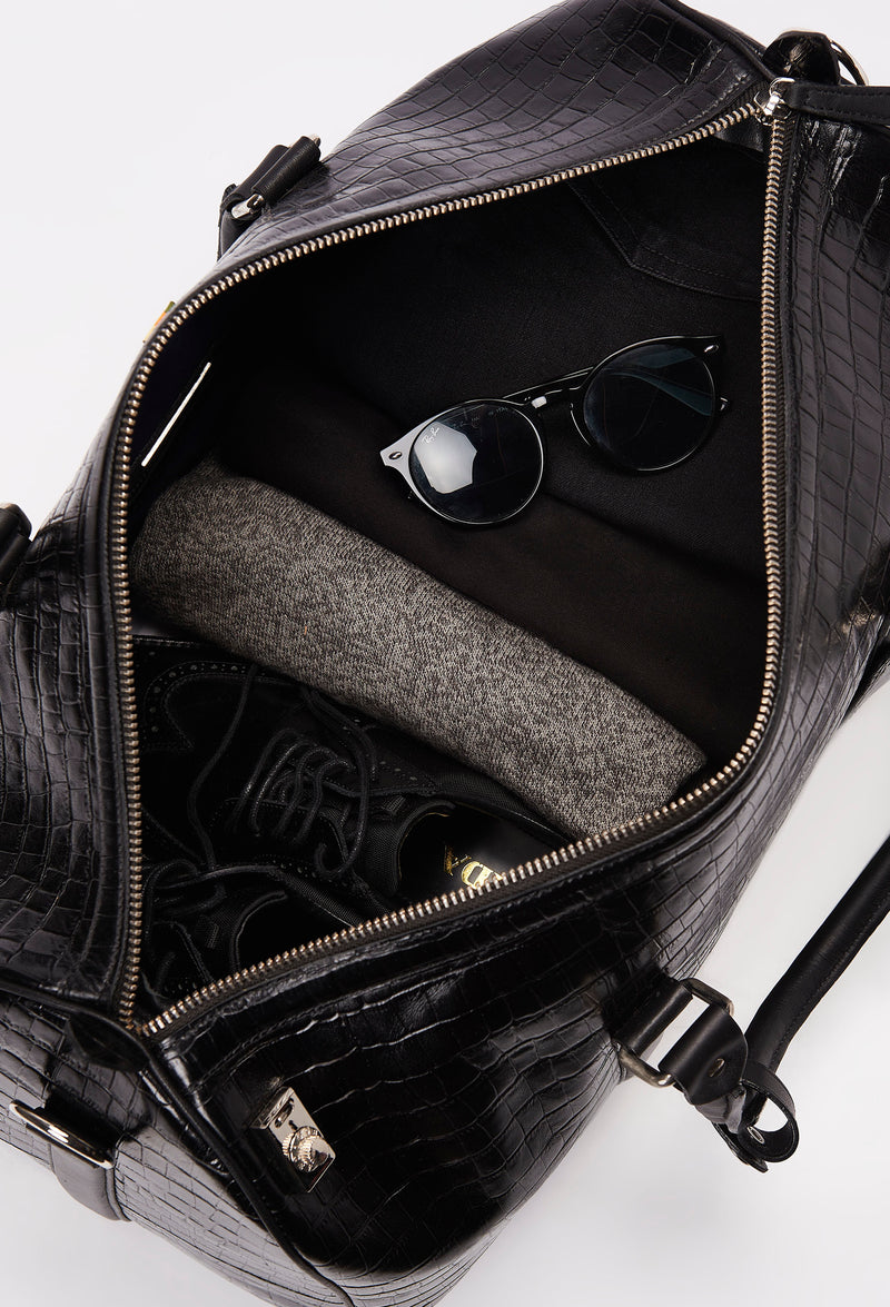 Interior of a Black Croco Leather Duffel Bag with lock closure packed with clothes, sunglasses and a pair of shoes.