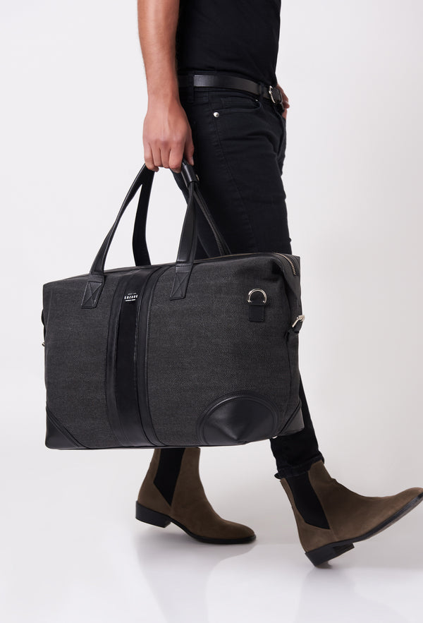 A model carries a sophisticated black large canvas duffel bag, showcasing its luxurious design. The bag features leather stripes on the front, adding to its elegant appeal. The model confidently displays the bag's size and craftsmanship while exuding a sense of style and sophistication.
