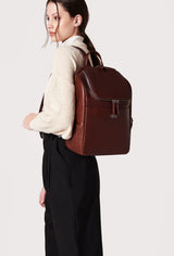 A model carries a sophisticated coffee leather backpack, showcasing its sophisticated design. The bag features unique needlework on its side, adding to its elegant appeal. The model confidently displays the bag's size and craftsmanship while exuding a sense of style and elegance.