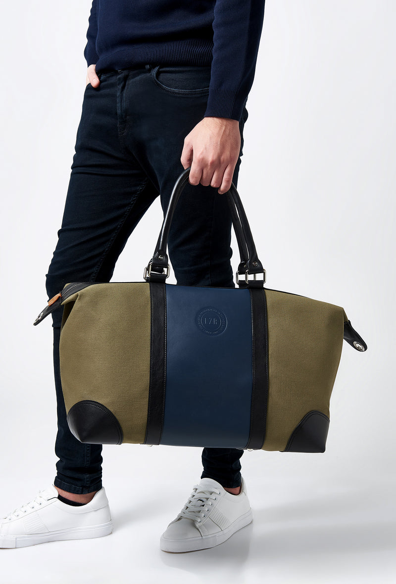 A model carries a sophisticated olive canvas and leather duffel bag, showcasing its luxurious design. The bag features a leather blue strap on the front, adding to its elegant appeal. The model confidently displays the bag's size and craftsmanship while exuding a sense of style and sophistication.
