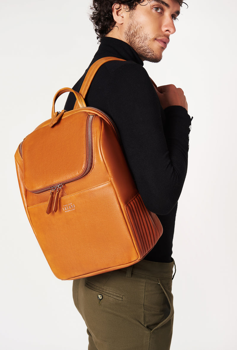 A model carries a sophisticated tan leather backpack, showcasing its sophisticated design. The bag features unique needlework on its side, adding to its elegant appeal. The model confidently displays the bag's size and craftsmanship while exuding a sense of style and elegance.