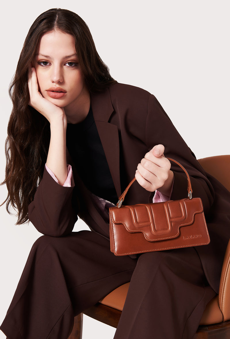 A model carries a sophisticated tan leather crossbody flap bag hilda, showcasing its sophisticated design. The bag features a raised design flap, adding to its elegant appeal. The model confidently displays the bag's size and craftsmanship while exuding a sense of style and elegance.