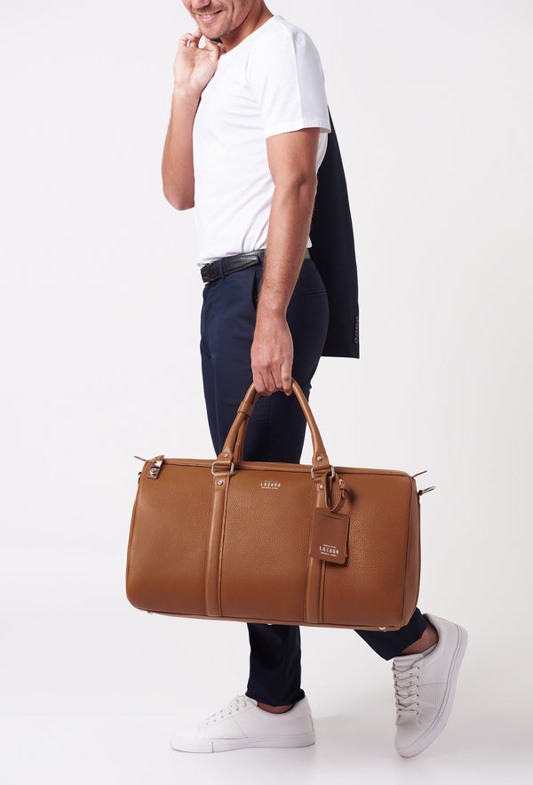 A model carries a sophisticated tan leather duffel bag, showcasing its luxurious design. The bag features a lock closure, adding to its elegant appeal. The model confidently displays the bag's size and craftsmanship while exuding a sense of style and sophistication.