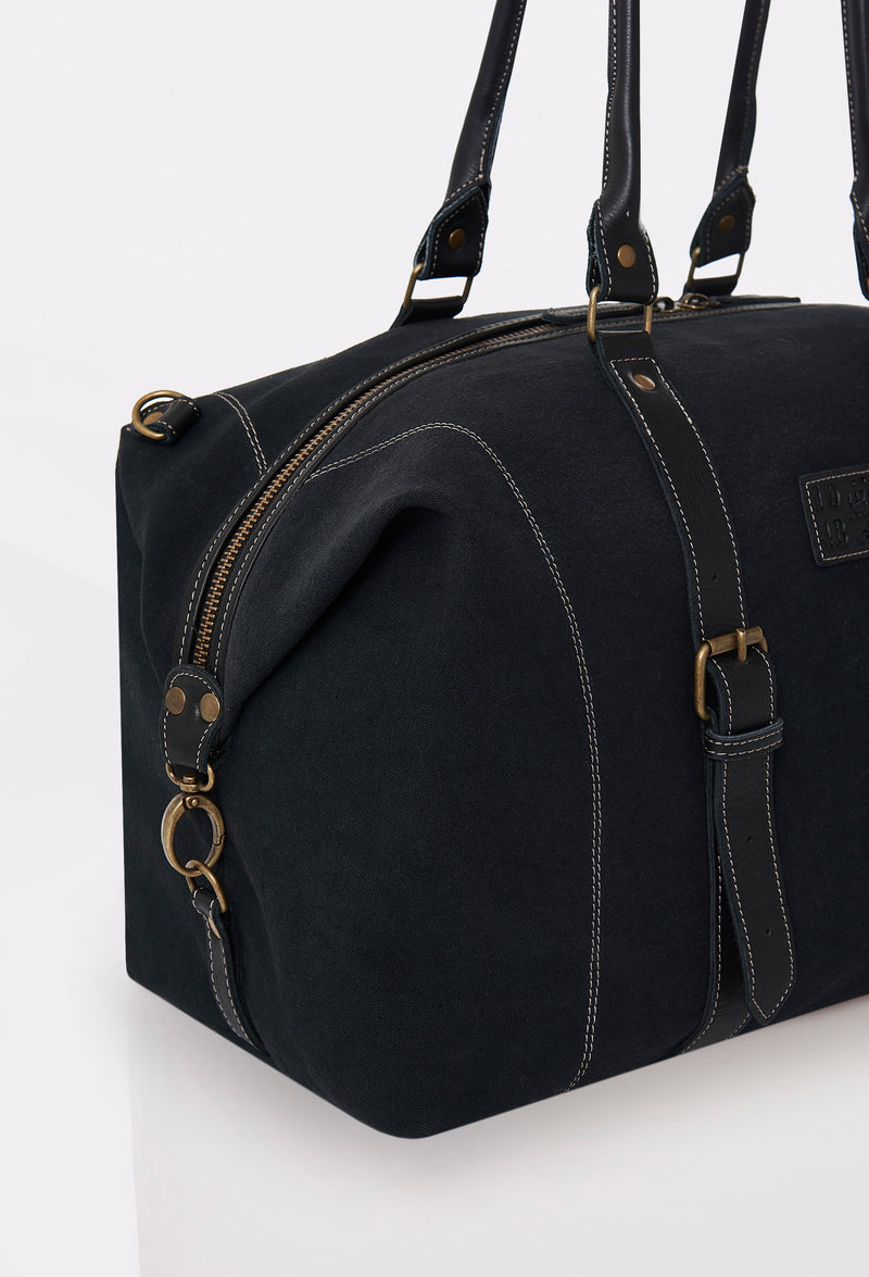 Partial photo of a Black Canvas Duffel Bag with leather handles, a zippered main compartment, and distressed antiqued bronze fittings.