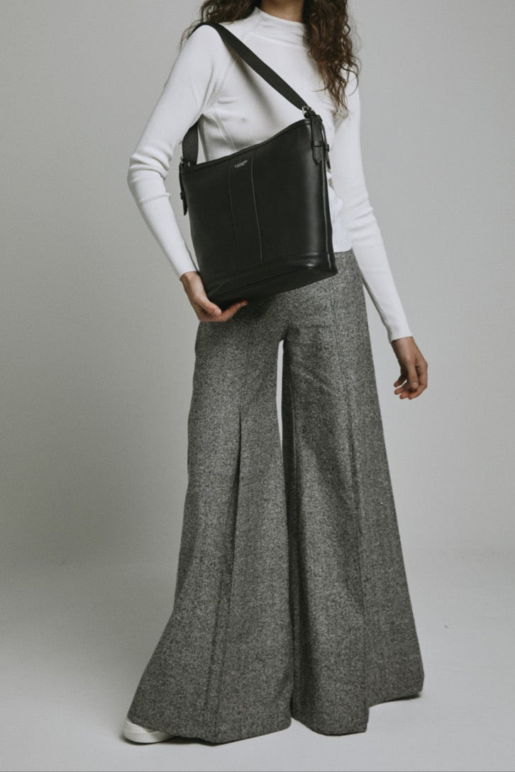 Woman holding a black bag wearing a white top and gray wide-leg pants on a plain background