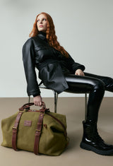 A stylish Olive Canvas Duffel Bag sits on the ground with a model seated showcasing its size and design.