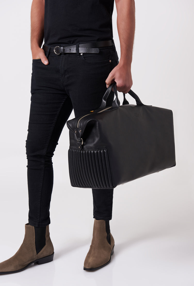 A model carries a sophisticated black leather duffel bag, showcasing its luxurious design. The bag features a unique needlework on its sides, adding to its elegant appeal. The model confidently displays the bag's size and craftsmanship while exuding a sense of style and sophistication.