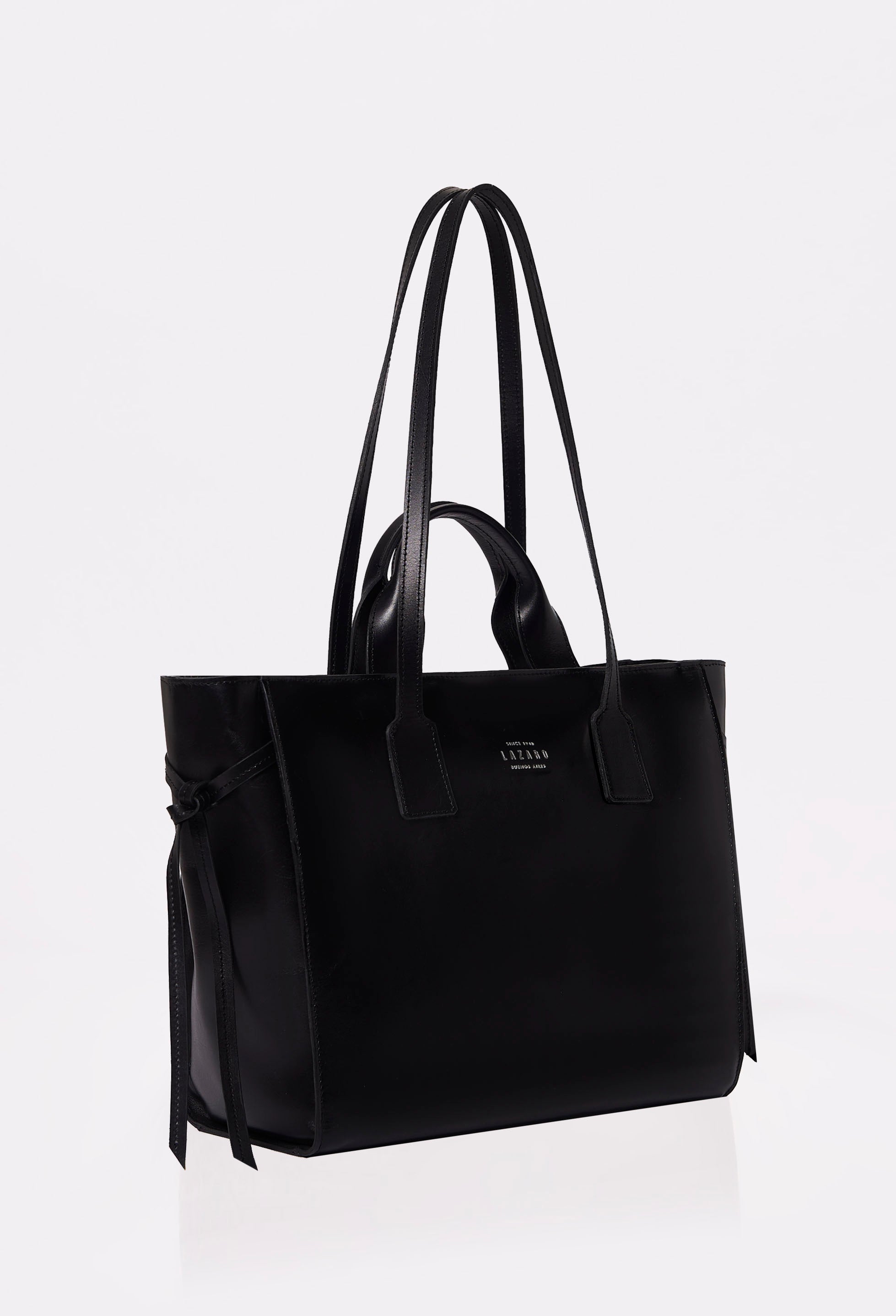 Side of a Black Leather Tote Bag Lambro with Lazaro logo, leather handle and straps and decorative knots on the sides.