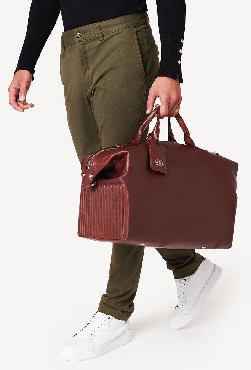 A model carries a sophisticated coffee leather duffel bag, showcasing its luxurious design. The bag features a unique needlework on its sides, adding to its elegant appeal. The model confidently displays the bag's size and craftsmanship while exuding a sense of style and sophistication.