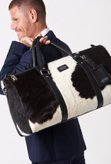 A model carries a sophisticated cowhide leather duffel bag on his back, showcasing its luxurious design. The bag features a prominent lock closure, adding to its elegant appeal. The model confidently displays the bag's size and craftsmanship while exuding a sense of style and sophistication.