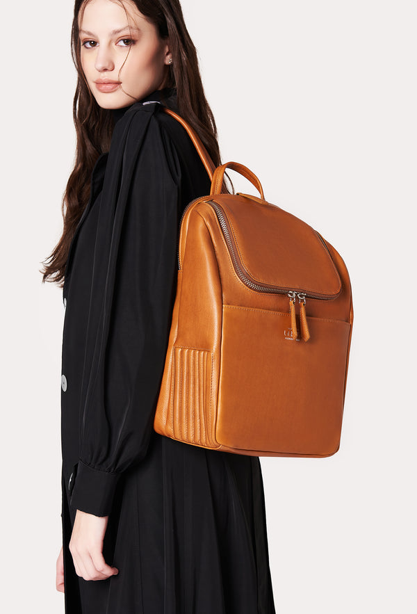 A model carries a sophisticated tan leather backpack, showcasing its sophisticated design. The bag features unique needlework on its side, adding to its elegant appeal. The model confidently displays the bag's size and craftsmanship while exuding a sense of style and elegance.
