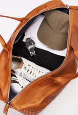 Interior of a Tan Leather Duffel Bag packed with a baseball cap, a watch, clothes and a pair of shoes.