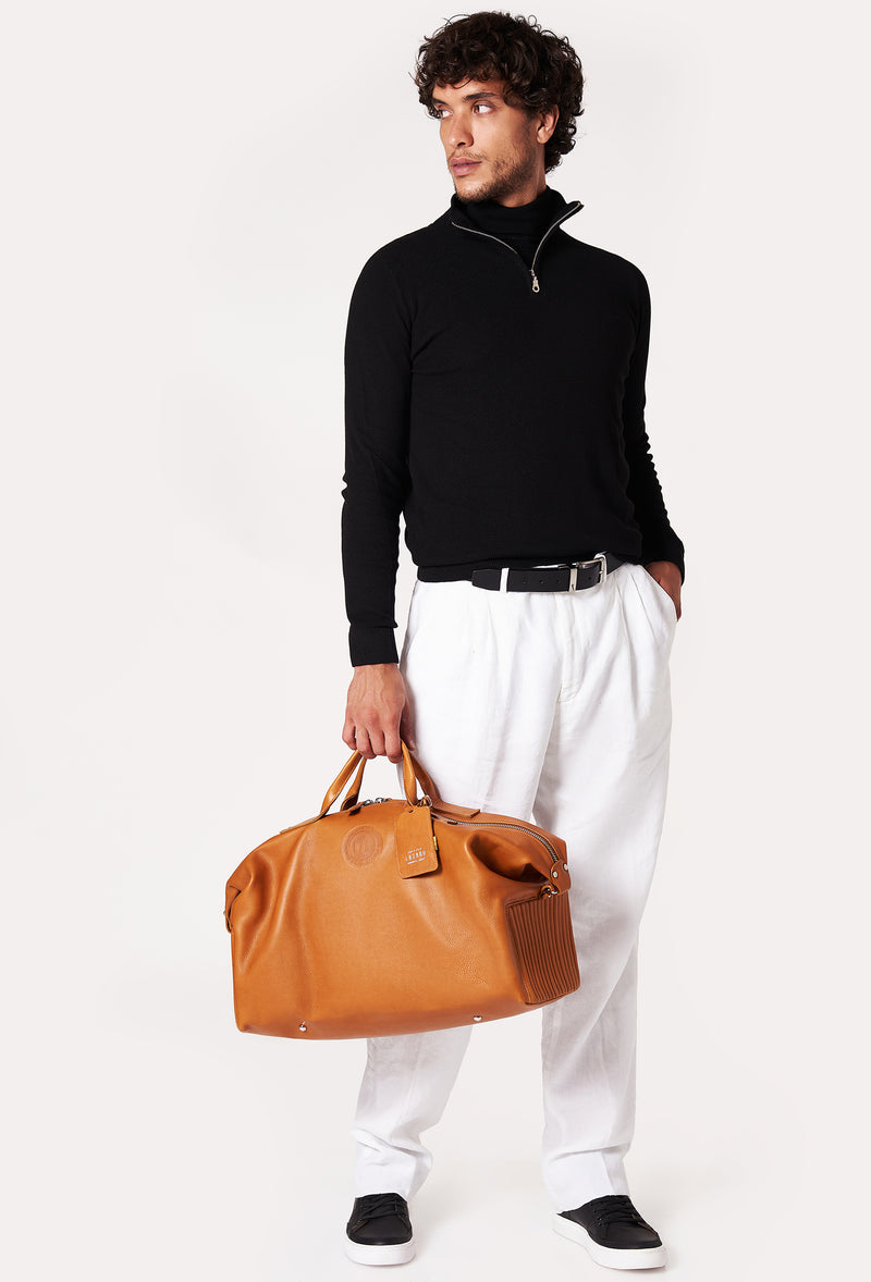 A model carries a sophisticated tan leather duffel bag, showcasing its luxurious design. The bag features a unique needlework on its sides, adding to its elegant appeal. The model confidently displays the bag's size and craftsmanship while exuding a sense of style and sophistication.