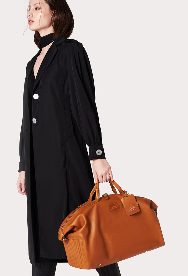 A model carries a sophisticated tan leather duffel bag, showcasing its luxurious design. The bag features a unique needlework on its sides, adding to its elegant appeal. The model confidently displays the bag's size and craftsmanship while exuding a sense of style and sophistication.